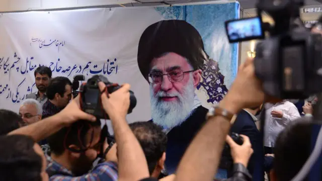 Ayatollah Khamenei shown on a white-cloth banner with Persian writing drawn on it. Cameras with video screens are pointed towards the banner.