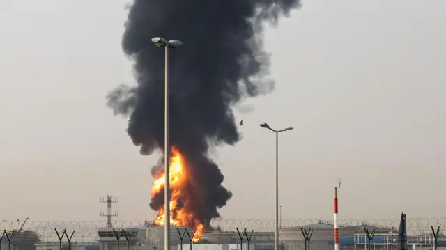 在阿拉伯联合大公国杜拜，杜拜国际机场附近的一处区域升起滚滚浓烟与烈焰。画面中可见数栋低矮建筑，四周环绕着一道保全围栏，似乎由盘绕的带刺铁丝或刀片刺网构成。