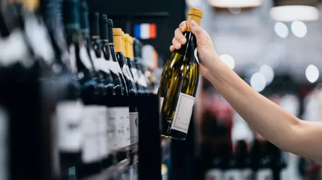 Egy kéz fehérboros üvegért nyúl egy szupermarket polcán.