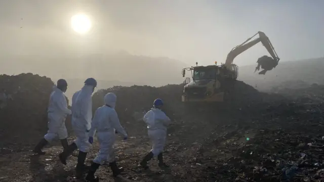 Agentes de la Policía de Essex registran un vertedero con overoles blancos, cascos y mascarillas. Al fondo se ve una excavadora con montones de residuos y un sol brumoso.