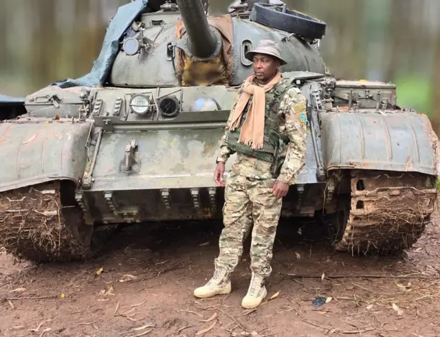 Willy Ngoma ari imbere y'igifaru cya gisirikare yambaye imyenda ya gisirikare n'ingofero