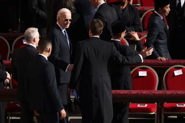 Joe Biden kırmızı koltuklar arasında, çevresinde başka erkekler var