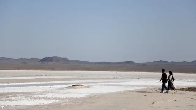 Un hombre y una mujer caminan por el lecho seco del lago salado Hoz-e Soltan en Qom: se puede ver sal en la superficie y montañas en la distancia.