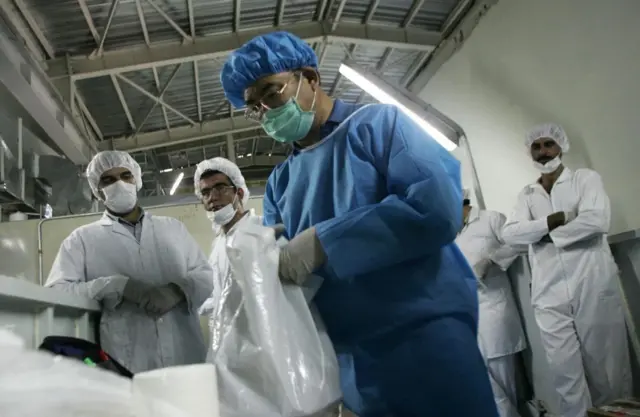 صورة أرشيفية بتاريخ 8 أغسطس/آب 2005، لفنيين إيرانيين ينظرون إلى مفتش من الوكالة الدولية للطاقة الذرية يعمل في منشآت تحويل اليورانيوم في أصفهان، على بعد 420 كيلومتراً جنوب طهران، ويرتدي مفتش الوكالة بدلة زرقاء وكمامة بيضاء، بينما يغطي رأسه بقبعة طبية زرقاء، ويرتدي الفنيون سترة بيضاء وكمامة وقبعة طبية بيضاء.