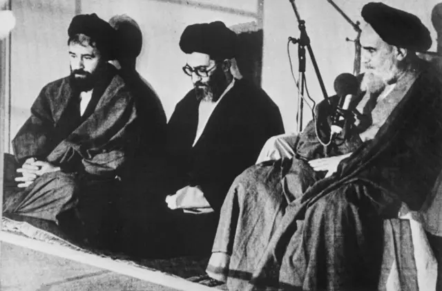 آیت الله خمینی در حال سخنرانی و آقای خامنه ای و احمد خمینی در کنارش روی زمین نشسته ‌اند و به او گوش می کنند