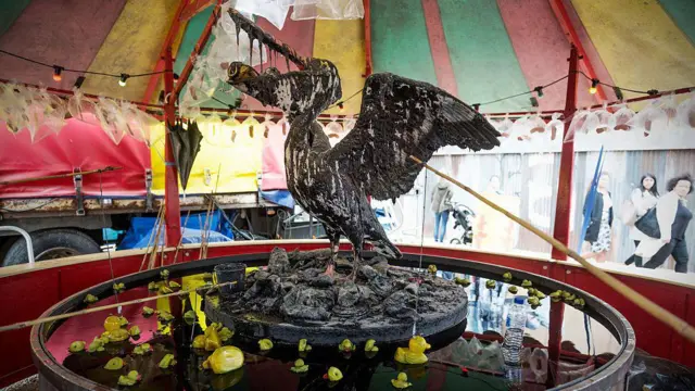 Традиційна ярмаркова гра hook-a-duck, але з несподіваним поворотом. Під кольоровим смугастим полотняним наметом великий білий морський птах сидить на каменях у центрі чана з темною нафтою, у якій він частково вимазаний. Його крила розпростерті, дзьоб відкритий, а всередині – зім’яті бляшанки від напоїв. Унизу – маленькі гумові качки, що перевернулися в мастилі. Ймовірно, це коментар щодо шкоди, яку нафтові розливи завдають екосистемам і дикій природі.