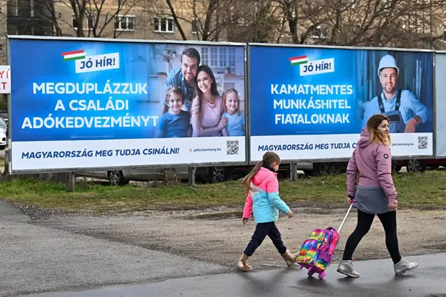Matka i córka idą chodnikiem przed dwoma billboardami. Na pierwszym widnieje czteroosobowa rodzina ze sloganem „podwajamy ulgi podatkowe dla rodzin”. Na drugim – pracownik w kasku ze sloganem „nieoprocentowane kredyty pracownicze dla młodych ludzi”.