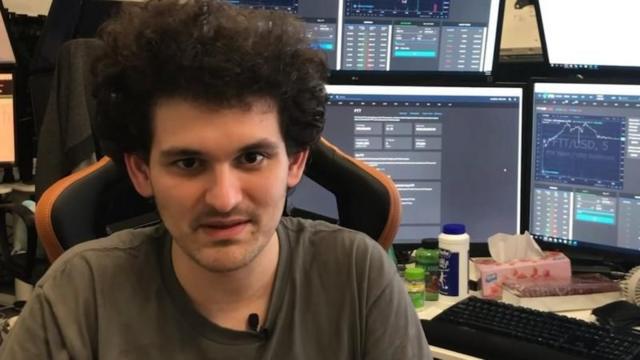 Sam em frente a monitores de computador mostrando gráficos