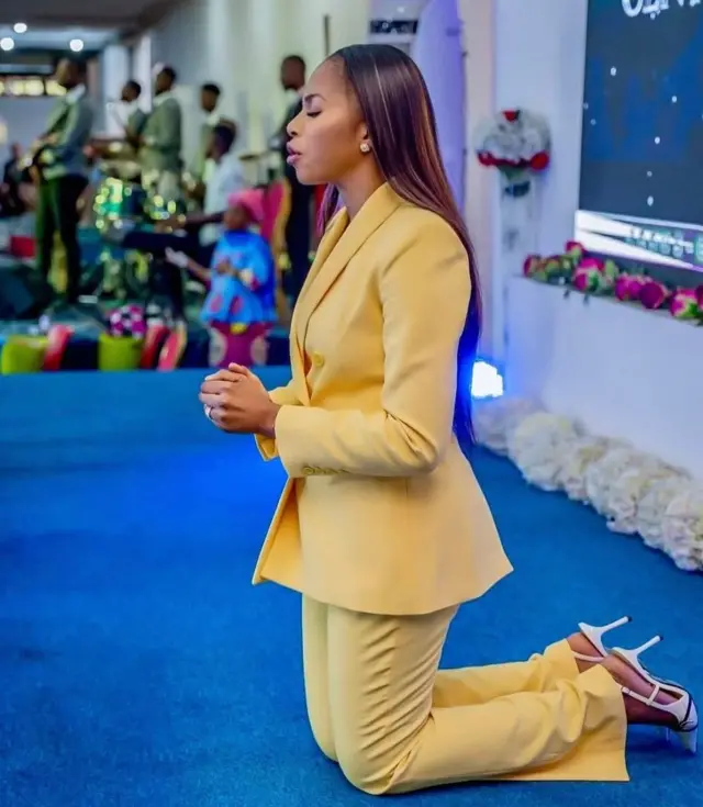 Grâce Kutino apfukamye arimo gusenga yambaye imyenda y'umuhondo inyuma mu ifoto harimo abaririmbyi