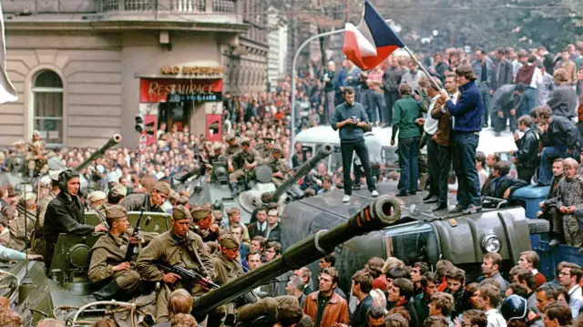 Людна демонстрації в Празі, кольорове фото. Кілька молодих людей стоять на даху автомобіля, тримаючи прапор Чехословаччини. Поруч у натовпі –танк з радянськими солдатами на ньому.