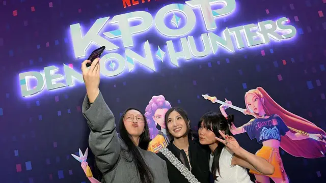 세 명의 여성이 사진을 찍고 있다. 왼쪽에 있는 여성은 손에 휴대폰을 들고 공중에 올린 채 셀카를 찍고 있으며, 다른 여성은 미소를 짓고 있고, 또 다른 여성은 평화의 손모양을 하고 있다. 배경에는 영화의 애니메이션 캐릭터들이 담긴 포스터와 함께 영화 제목이 굵고 흰 글씨로 적혀 있어 눈길을 끈다.