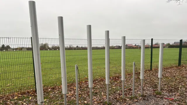La imagen muestra una instalación musical al aire libre compuesta por siete tubos metálicos cilíndricos y altos dispuestos en fila a lo largo de un camino de tierra.