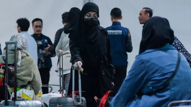 Seorang WNI perempuan dari Iran dengan pakaian serba tertutup hijab dan masker sedang mendorong sebuah koper di antara kerumunan orang. Ia tampak jalan dengan menunduk.