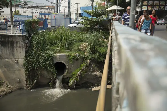 Pessoas atravessando ponte; abaixo, é possível ver grande tubulação depositando esgoto