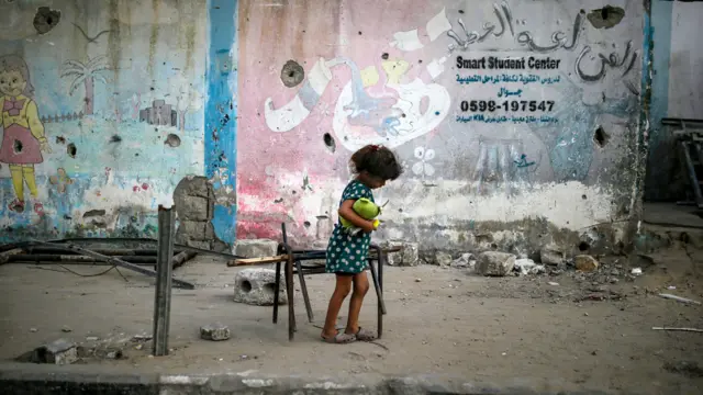 Uma criança caminha com o que parece ser um boneco na mão por meio de escombros. Ela está de cabeça baixa e sozinha.