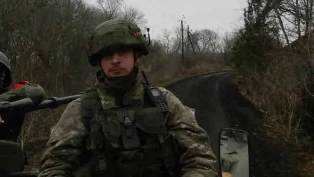 To‘liq qurollangan askar, tank ustida o‘tirib, barglari to‘kilgan daraxtli yo‘l bo‘ylab harakatlyapti.