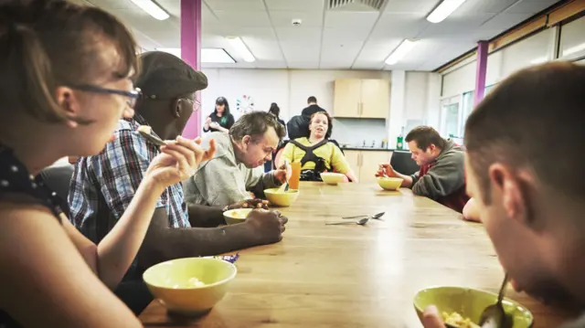 Seis pessoas se alimentam sentadas ao redor de uma mesa de uma instituição de caridade.