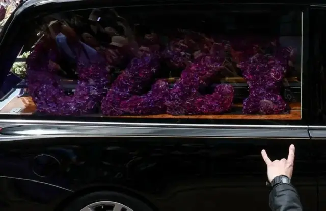 В окне машины видна выложенная цветами надпись Ozzy. Каждая буква как отдельный венок. Вероятно, за ними находится гроб.