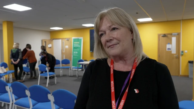 Mujer rubia con flequillo, con un jersey negro y un cordón rojo para visitas, con la cabeza ligeramente ladeada. De pie frente a una sala de reuniones pintada de amarillo, con sillas azules dispuestas en círculo.