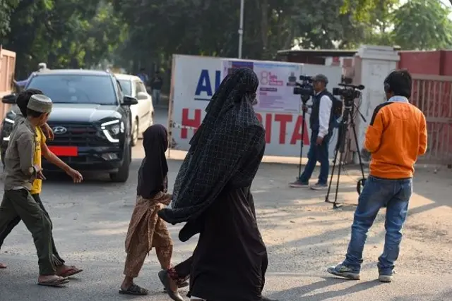 Bir kadın ve kız çocuğu odakta, arka planda Al Falah Hospital girişi görünüyor. Bir kameraman burada çekim yapıyor. 