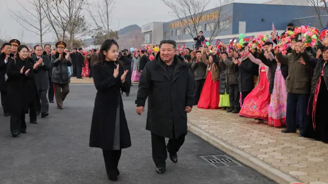 Młoda Koreanka z Północy w ciemnym płaszczu idzie obok Koreańczyka z Północy w średnim wieku. Są witani przez tłum.
