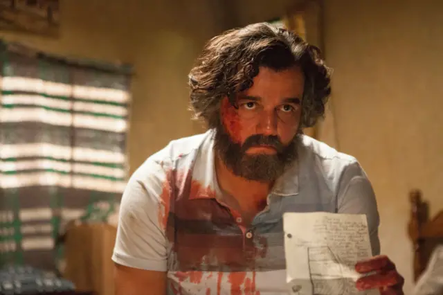 O ator Wagner Moura aparece como Pablo Escobar, com o rosto e a camisa manchados de sangue, segurando um papel com anotações enquanto encara a câmera com expressão tensa em cena da série Narcos.