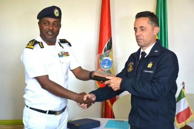 ጻዕዳ ወተሃደራዊ ልብሲ ሓይሊ ባሕሪ ዝተኸደነ ኣዛዚ ሓይሊ ባሕሪ ኢትዮጵያ ምስ ሰማያዊ ልብሲ ዝተኸደነ መራሒ ጉጅለ ሓይሊ ባሕሪ ኢጣልያ ሰላምታ እናተለዋወጠ። ባንዴራ ክልቲአን ሃገራት ድማ ኣብ ድሕረባይታኦም ኣለዋ።