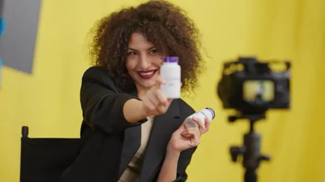 Mulher em frente a uma câmera promove produtos em frascos em suas mãos