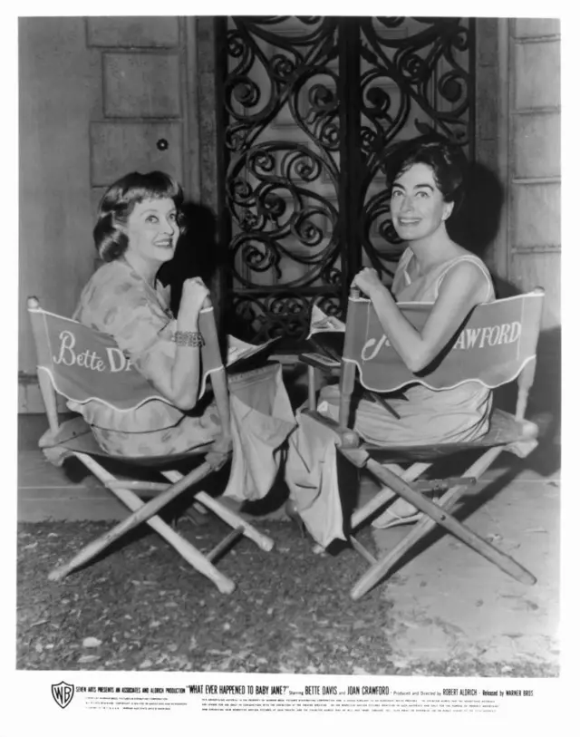 Foto em preto e branco de Bette Davis e Joan Crawford, sentadas em cadeiras de diretor com seus nomes bordados nas costas