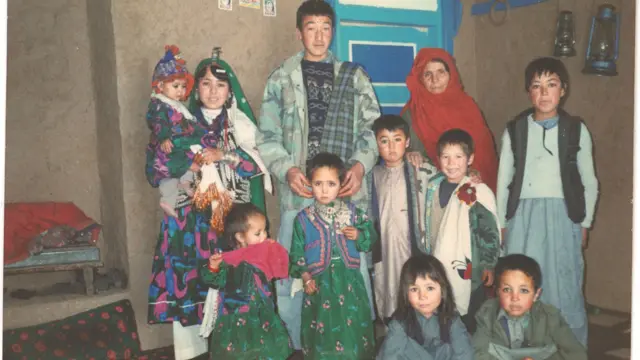 Zahra's wider family inside a house, wearing traditional Afghan clothes. She squats at the front, with dark hair and a fringe, next to her young brother who has cropped hair. Five other young children stand behind her – the girls in green and pink smocks over white trousers. Her mother is at the back wearing a red headscarf.