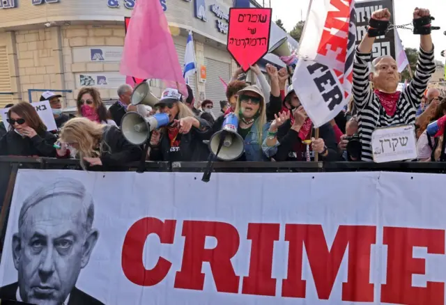 Manifestantes en Jerusalén con megáfonos y una pancarta que muestra una imagen de Netanyahu con la palabra "crimen" 
