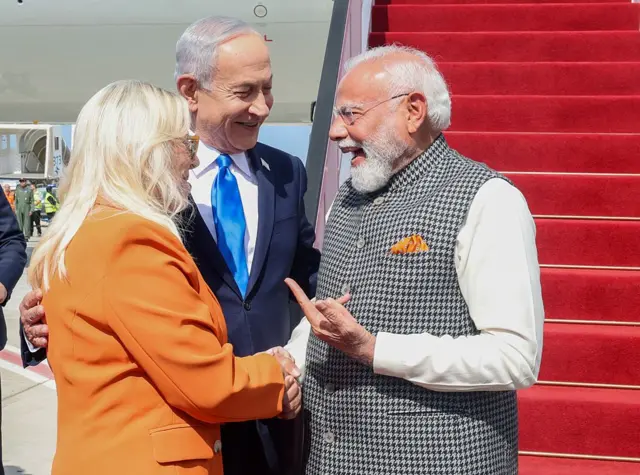 Modi, Netanyahu ve turuncu ceketli bir kadın gülerek sohbet ediyorlar. Arka planda bir uçağın kırmızı halıyla kaplı merdivenleri görünüyor. 