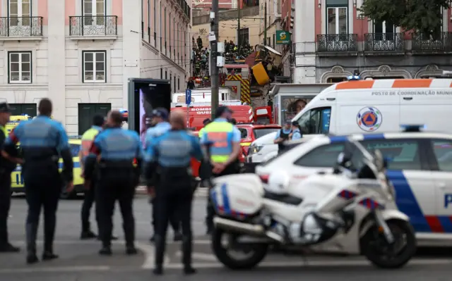 Policjanci stoją w pobliżu miejsca wypadku, na którym widać przewróconą kolejkę linowo-terenową rozbitą na ulicy