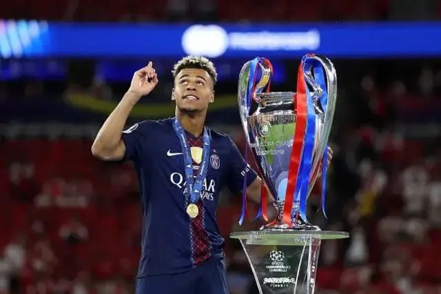 Désiré Doué afashe ku gikombe cya Champions League, areba hejuru. Yambaye ikabutura n'umupira w'amaboko magufi byiganjemo ubururu bwijimye, yambaye n'umudari mu ijosi. 