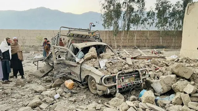 Bir kamyonet yıkıntılar arasında kalmış. Yanında aracın haline bakan üç kişi var.