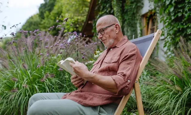 Un homme d'age mûr savoure un moment de calme en lisant un livre, allongé sur une chaise longue pliante dans un jardin luxuriant.
