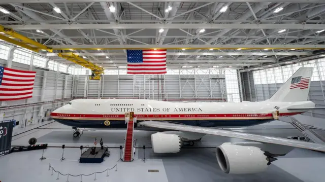 full shot of new Air Force One plane in a grey hangar. Two large American flags hang in the background.