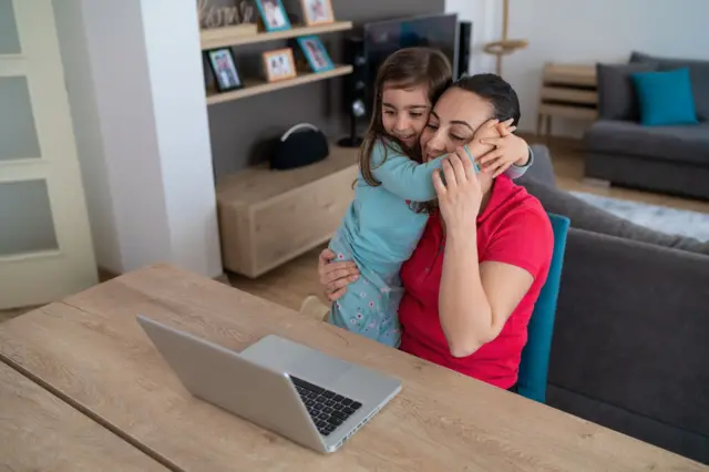 امرأة تجلس إلى طاولة أمام حاسوب محمول، تحتضنها طفلة صغيرة وتلفّ ذراعيها حول عنقها