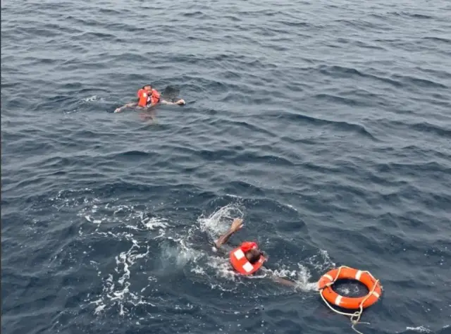 شخصان يرتديان ملابس إنقاذ برتقالية اللون، وسط البحر. الصورة التقطت نهاراً والبحر فيه أمواج منخفضة العلو.