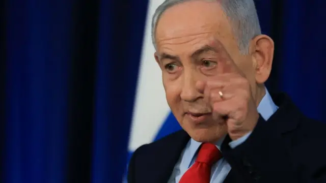 Benjamin Netanyahu de lado, con una mano en algo, durante una rueda de prensa. Viste traje oscuro, camisa azul claro y corbata roja.