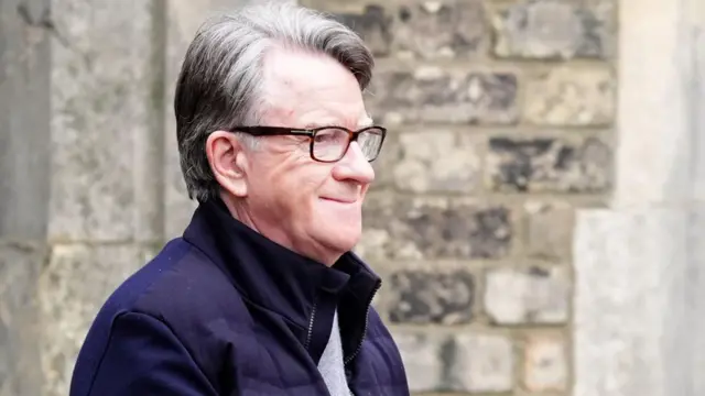 Peter Mandelson w szarej bluzie, granatowej kurtce i okularach uśmiecha się, gdy policja wyprowadza go po aresztowaniu na początku tego tygodnia