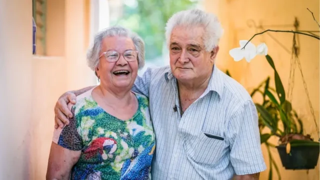 Mulher idosa, cabelo brancos e usando óculos, abraça homem idoso,cabelos brancos, com camisa