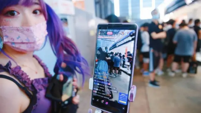 Jovemaposta ganha inter de milãocabelo roxo e máscara segurando celular mostrando live