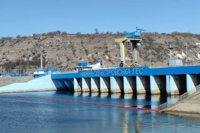 Часть гидротехнического сооружения с надписью «Нижнеднестровская ГЭС» на синей панели. Перед дамбой в воде виден установленный красный плавучий барьер, вероятно предназначенный для сдерживания загрязнений. На заднем плане — холм с сухой растительностью и отдельными постройками. Небо ясное, погода солнечная.