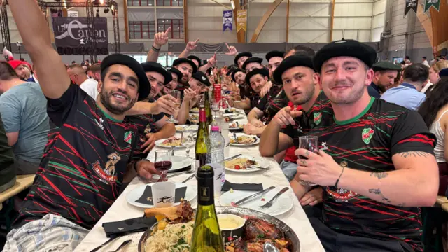 Egy csoport fiatal férfi ül egy hosszú, étellel és borral megrakott asztal mellett, azonos sportmezben és svájcisapkában