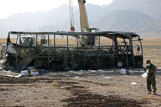 بقایای اتوبوس سوخته و یک نیروی امنیتی طالبان ایستاده در کنارش 