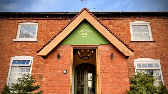 Foto exterior del Fox Inn. Es un edificio de dos plantas, de ladrillo rojo, con cuatro ventanas con marcos blancos visibles. El porche delantero tiene un letrero verde sobre la puerta que dice "El Zorro, Ryton" con la ilustración de un zorro corriendo. A ambos lados del porche hay dos árboles de Navidad.