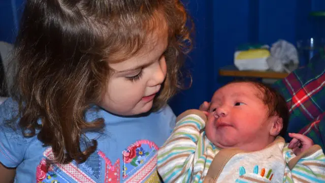 Kate patrzy w dół na noworodka Jamesa trzymanego w ubraniu o wzorze.