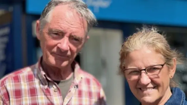 トミー氏は赤と白と黄色のチェックシャツ、シャーリー氏はデニムシャツを着て、笑顔で並んで立っている