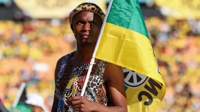 Um homem negro vestindo uma camiseta regataroleta com duas bolasleopardo, carrega uma bandeira verde e amarela com as iniciais ANC, do Congresso Nacional Africano